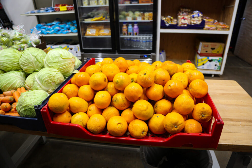 a table with a flat of oranges and flat of cabbage with coolers of drinks and shelves of food in background